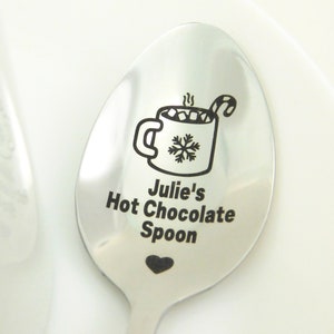 May include: A silver spoon with a personalized inscription that reads "Julie's Hot Chocolate Spoon". The spoon features an engraved design of a mug of hot chocolate with marshmallows, a candy cane, and a snowflake.