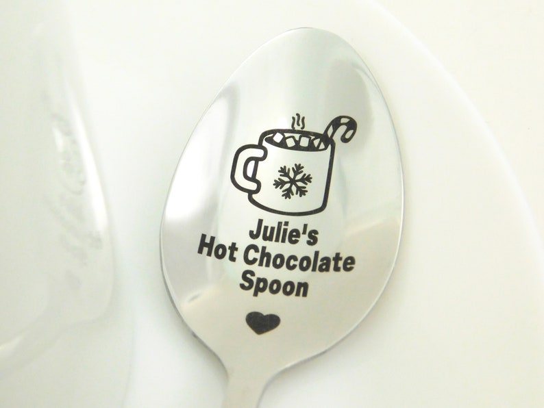 May include: A silver spoon with a personalized inscription that reads "Julie's Hot Chocolate Spoon". The spoon features an engraved design of a mug of hot chocolate with marshmallows, a candy cane, and a snowflake.