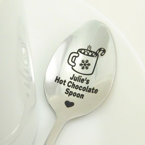 May include: A stainless steel spoon with a black engraved design of a mug of hot chocolate with marshmallows and a candy cane. The spoon is engraved with the text "Julie's Hot Chocolate Spoon" and a small heart.