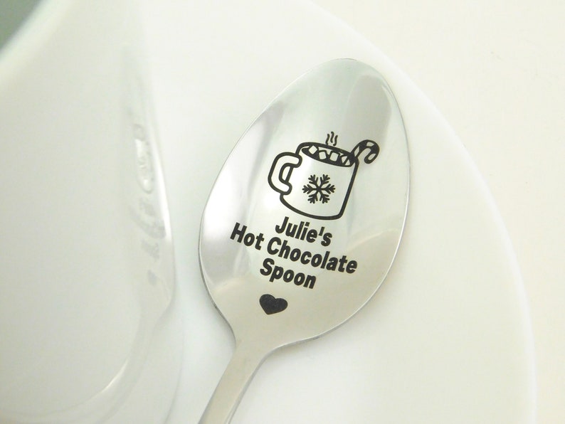 May include: A stainless steel spoon with a black engraved design of a mug of hot chocolate with marshmallows and a candy cane. The spoon is engraved with the text "Julie's Hot Chocolate Spoon" and a small heart.