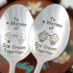 Puede incluir: Dos cucharas de plata con el texto "Para toda la vida de helado juntos" grabado en ellas. Cada cuchara presenta un cono de helado de dibujos animados y una paleta con corazones.