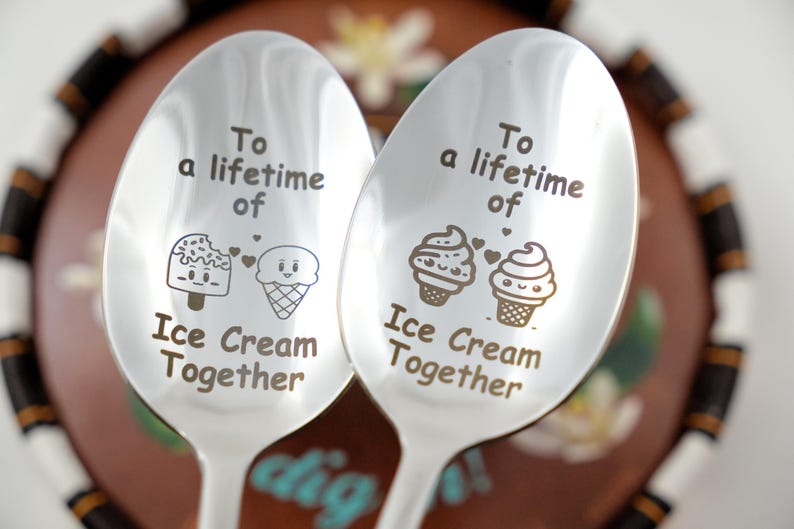Puede incluir: Dos cucharas de plata con el texto "Para toda la vida de helado juntos" grabado en ellas. Cada cuchara presenta un cono de helado de dibujos animados y una paleta con corazones.