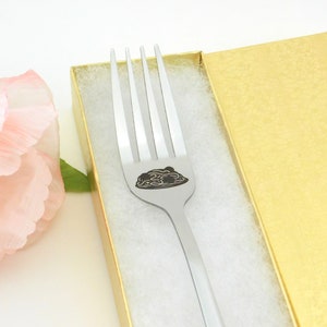 May include: A silver fork with a black engraved design of a plate of spaghetti and meatballs. The fork is in a white and gold gift box.