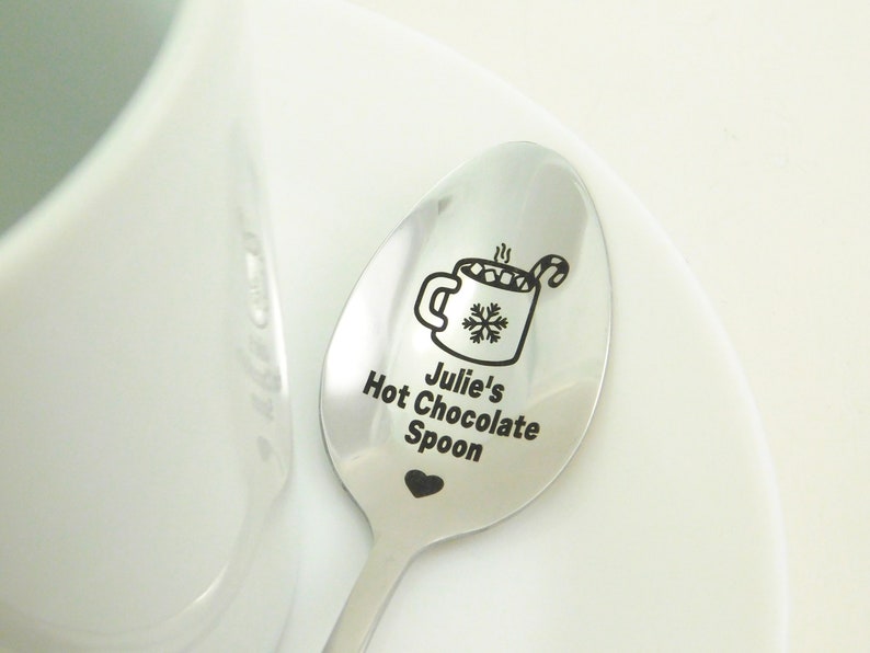 May include: A silver spoon with a black engraved design of a mug of hot chocolate with marshmallows and a candy cane. The text "Julie's Hot Chocolate Spoon" is engraved below the mug.