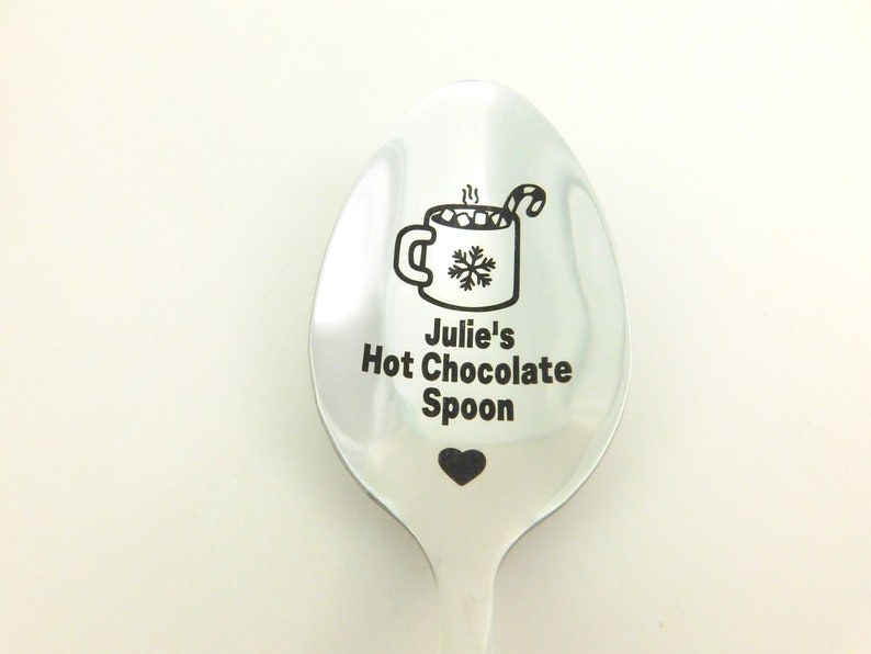 May include: A silver spoon with a black engraved design of a mug of hot chocolate with marshmallows and a candy cane, along with the text "Julie's Hot Chocolate Spoon" and a small heart.