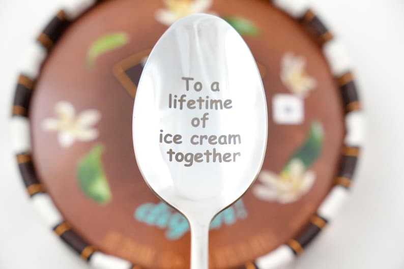 Puede incluir: Una cuchara de plata con la inscripci&oacute;n "To a lifetime of ice cream together" grabada en ella. La cuchara est&aacute; enfocada, y un plato de postre marr&oacute;n y blanco est&aacute; desenfocado en el fondo.