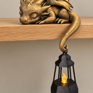 May include: A gold-colored sleeping dragon figurine with textured scales and wings, resting on a ledge. Its long, curved tail hangs down, holding a black hexagonal lantern that contains a small, glowing yellow LED flame light.