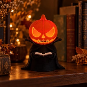 May include: A glowing orange jack-o'-lantern figure with a carved, menacing face, wearing a black cape with a high collar. The figure holds a small, open white book in its hands, creating a light-up decorative piece with a dark, eerie aesthetic.