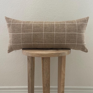 May include: A rectangular lumbar pillow featuring a textured tan fabric with a white grid pattern. The pillow has a visible zipper on the lower left side and is positioned horizontally, showcasing a woven appearance with clean, straight lines.