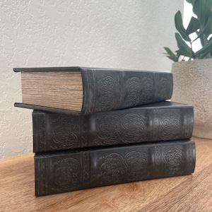 May include: Three black leather-bound books stacked on top of each other. The books have an intricate embossed design and are on a wooden surface.