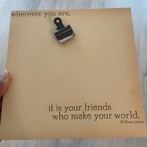 May include: A square, tan-colored memo board with a metal clip at the top. The board features the quote, "wherever you are, it is your friends who make your world." The quote is attributed to William James.