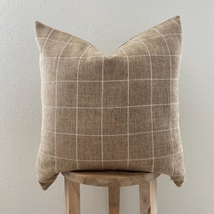 May include: A square decorative pillow with a brown plaid pattern. The pillow is made of a textured fabric and has a grid design in white. The pillow is resting on a wooden stool.