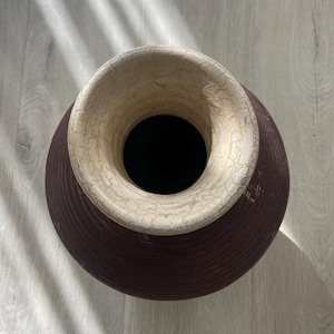 May include: A large, decorative vase with a wide, circular opening. The vase has a dark burgundy body with a textured, ridged surface. The rim is a light beige color with a cracked, aged appearance. The interior is black.