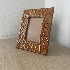 May include: A rectangular picture frame with a glossy, mustard-brown finish and a raised, geometric lattice pattern covering the entire border. The frame features a centered, plain tan cardboard insert and is angled to show its depth and structure.