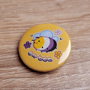 May include: A yellow button featuring a cartoon bee with purple, white and yellow stripes. The bee is surrounded by small yellow flowers and the text "em-bee".