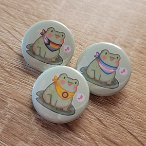 Peut inclure: Trois boutons ronds de grenouille verte avec des bandanas de couleurs différentes. La première grenouille porte un bandana arc-en-ciel, la deuxième grenouille porte un bandana de fierté transgenre et la troisième grenouille porte un bandana jaune avec un cercle noir.