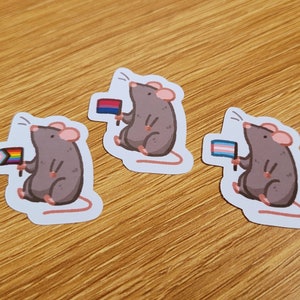 May include: Three stickers of cartoon rats, each holding a pride flag. The rats are grey with pink ears and feet. The flags represent different LGBTQ+ identities: bisexual, transgender, and the progress pride flag.