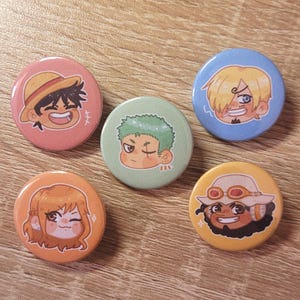 May include: Six round pins featuring cartoon characters from the anime One Piece. The characters are Luffy, Zoro, Nami, Sanji, Chopper, and Usopp. The pins are colorful and have a glossy finish.