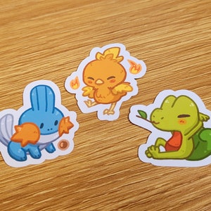 Peut inclure: Trois autocollants de personnages de dessins animés. Un personnage bleu et orange avec une queue, un personnage orange et jaune ressemblant à un oiseau avec des flammes, et un personnage vert et orange ressemblant à un lézard avec une feuille.