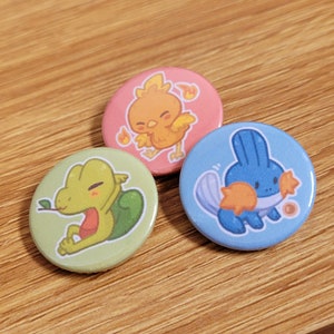 May include: Three round pins featuring cartoon images of Pokemon characters. The first pin is pink with a yellow bird with orange flames. The second pin is green with a green lizard with a brown leaf in its mouth. The third pin is blue with a blue and orange water Pokemon.