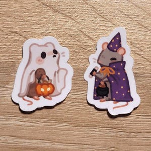 May include: Two Halloween-themed stickers. One is a white ghost holding a pumpkin. The other is a gray mouse wearing a purple cape with white polka dots and a matching hat, holding a cauldron. The stickers are on a wooden surface.