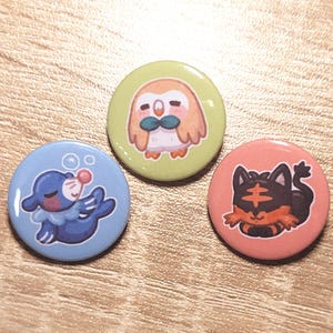 May include: Three circular pin badges featuring cartoon animal illustrations. One is light blue with a seal, another is light green with an owl, and the third is coral with a tiger-like cat.