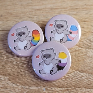 May include: Three round button pins with a cartoon raccoon design. Each raccoon has a different colored tail representing a pride flag: rainbow, transgender, and bisexual.