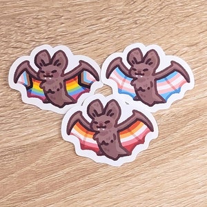 Może przedstawiać: Trzy brązowe naklejki z nietoperzami z tęczowymi i transgenderowymi flagami dumy na skrzydłach.