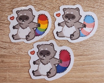 May include: Three sticker designs featuring a cartoon raccoon with a rainbow, bisexual, and transgender pride flag tail. Each sticker has a small red heart in the top left corner.
