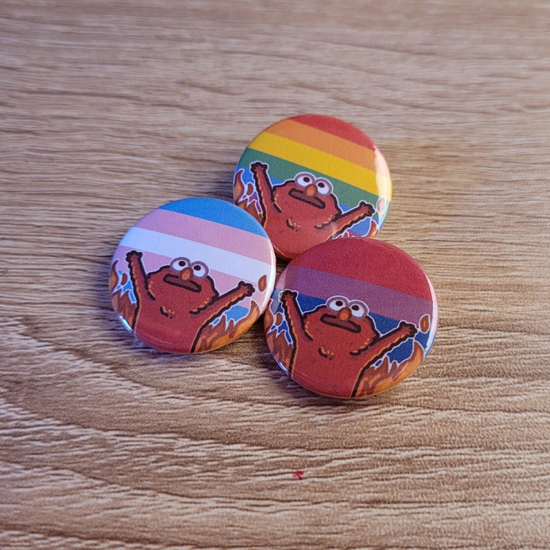 Peut inclure: Trois &eacute;pingles rondes en forme de bouton repr&eacute;sentant un personnage de dessin anim&eacute; rouge avec une expression surprise. Le personnage est sur un fond de rayures de diff&eacute;rentes couleurs. La premi&egrave;re &eacute;pingle a un fond de rayures arc-en-ciel, la deuxi&egrave;me a un fond de drapeau de fiert&eacute; transgenre et la troisi&egrave;me a un fond de drapeau de fiert&eacute; bisexuel.