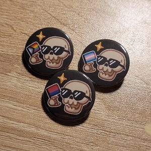 Może przedstawiać: Trzy czarne, okrągłe guziki z czaszką w okularach przeciwsłonecznych i flagą. Każdy guzik ma inny wzór flagi: tęczowy, transpłciowy i biseksualny. Na każdym guziku znajduje się również żółta gwiazda.