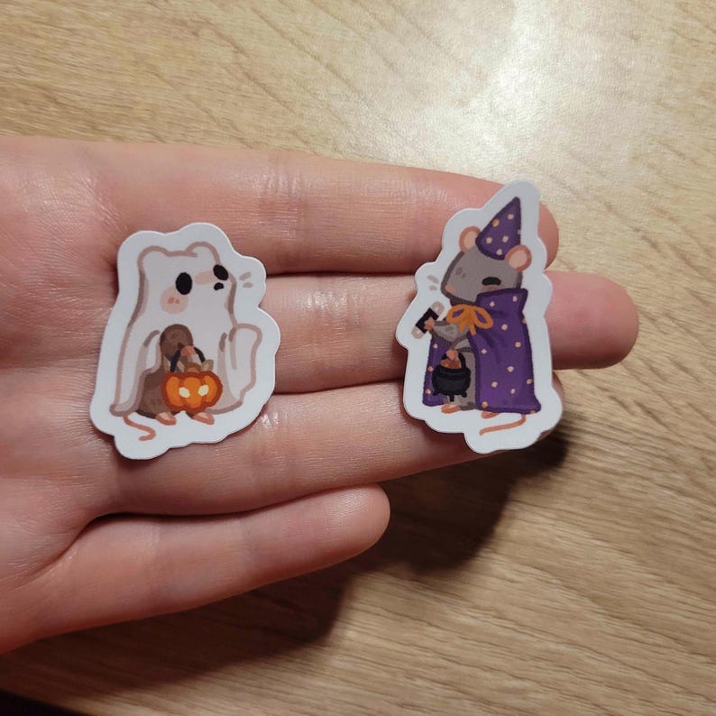 May include: Two Halloween-themed stickers. One sticker features a white ghost holding a pumpkin. The other sticker depicts a mouse in a purple wizard's cloak and hat, holding a cauldron. Both stickers have a white border.