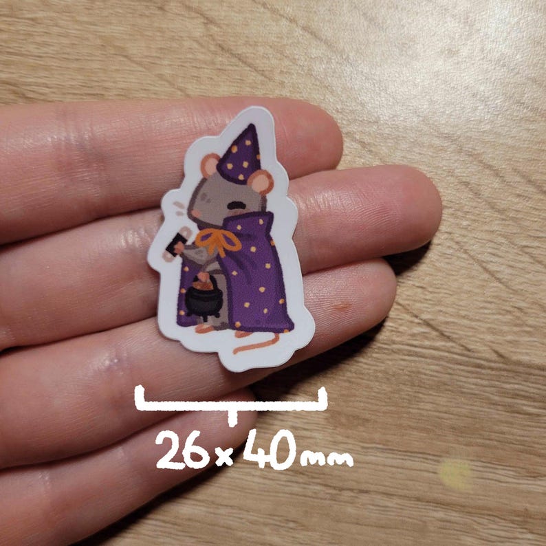 May include: A sticker featuring a cartoon mouse dressed as a wizard, wearing a purple cape with gold polka dots and a matching hat. The mouse holds a small cauldron and a wand. The sticker measures 26 x 40 mm.
