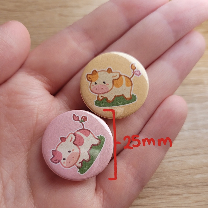 May include: Two round, 25mm diameter badges with cartoon cow designs. One badge has a light yellow background with a standing cow, the other a pink background with a lying cow. Both cows have pink and brown spots.