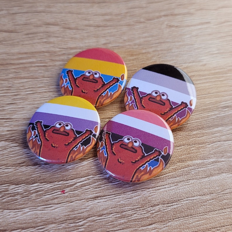Peut inclure: Quatre badges ronds avec diff&eacute;rents motifs de drapeaux arc-en-ciel. Chaque badge pr&eacute;sente un personnage de dessin anim&eacute; rouge, orange, jaune et brun avec une expression surprise. Le personnage est entour&eacute; de flammes. Les drapeaux sont : jaune, rouge et bleu ; noir, blanc et gris ; violet, blanc et rose ; et jaune, blanc et violet.