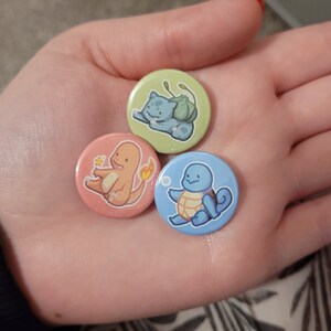 May include: Three round buttons featuring cartoon images of Pokemon characters. The buttons are green, pink, and blue. The green button features Bulbasaur, the pink button features Charmander, and the blue button features Squirtle.