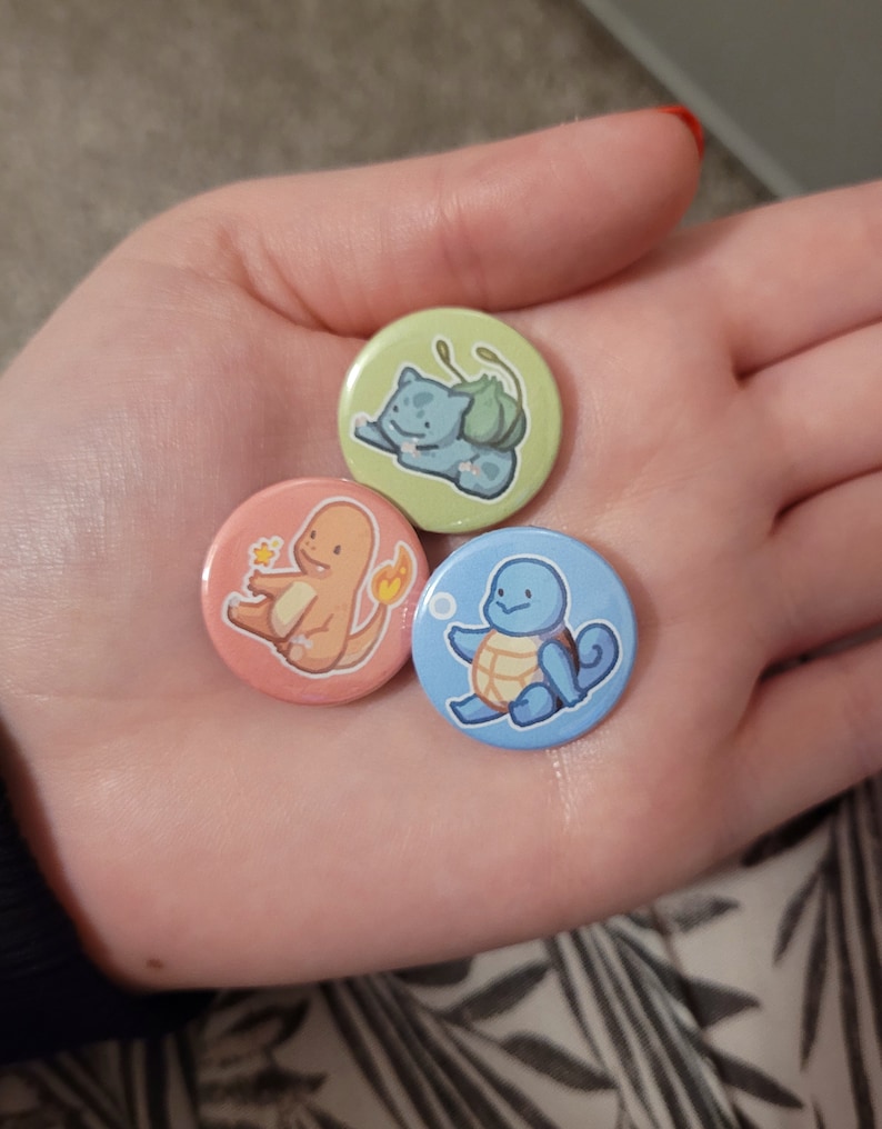 May include: Three round buttons featuring cartoon images of Pokemon characters. The buttons are green, pink, and blue. The green button features Bulbasaur, the pink button features Charmander, and the blue button features Squirtle.