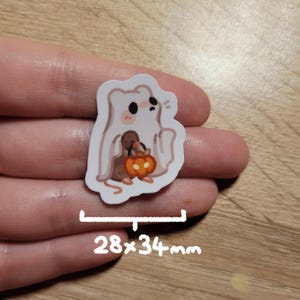 May include: A Halloween-themed sticker featuring a cartoon ghost holding a pumpkin-shaped bag. The sticker is white with a brown and orange pumpkin and measures 28 x 34 mm.
