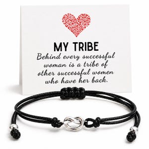 May include: A black cord bracelet featuring a central silver-toned metal knot charm and two silver-toned beads at the ends. Behind the bracelet is a white card with a red heart graphic and the text: MY TRIBE Behind every successful woman is a tribe of other successful women who have her back.