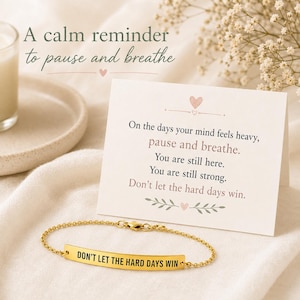 May include: A gold-toned metal bar bracelet with a delicate chain, featuring the engraved text 'DON'T LET THE HARD DAYS WIN'. It is displayed alongside a white card printed with the message: 'On the days your mind feels heavy, pause and breathe. You are still here. You are still strong. Don't let the hard days win.'