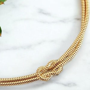 May include: A gold-toned necklace with a knot design. The necklace is made of a thin, braided chain.