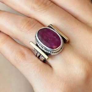 May include: A silver ring featuring a large oval ruby gemstone. The ring has gold-coloured accents on the sides. The ruby is a deep red colour, and the ring is worn on a hand with red nail polish.