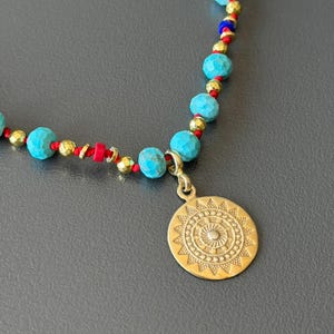 May include: A necklace featuring turquoise-colored beads, gold-colored beads, and a gold-colored pendant. The pendant has a sunburst design. The beads are strung on a red cord.