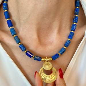 Può includere: Una collana con perline blu in lapislazzuli con dettagli dorati. La collana ha un ciondolo dorato con un design circolare. La collana è indossata.