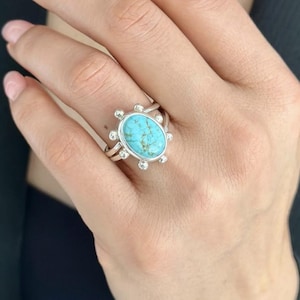 Puede incluir: Un anillo de plata con una piedra ovalada turquesa engastada en un engaste de plata decorativo. El anillo tiene un diseño delicado e intrincado.