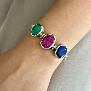 May include: Silver bracelet with three oval gemstones in green, magenta, and blue. The gemstones are set in individual silver bezels and linked together. The bracelet is worn on a wrist.