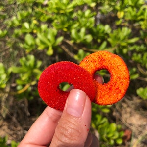 May include: Two red and orange ring-shaped candies with a grainy texture. The candies are held in a hand.