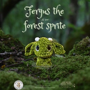 Może przedstawiać: Zielony, szydełkowy leśny skrzat o imieniu Fergus z białymi rogami i dużymi oczami. Skrzat siedzi na mchu. Tekst "Fergus the forest sprite -No sew- " jest napisany nad skrzatem.