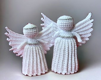 May include: Two white crocheted angels with silver trim. The larger angel is standing behind the smaller angel. Both angels have wings and are facing the right side of the image. The image is captioned with the text "@Monasheklekrok".