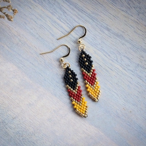 May include: A pair of dangle earrings with gold hooks and beaded drops. The beads are black, red, and yellow, arranged in a pattern resembling a flag.